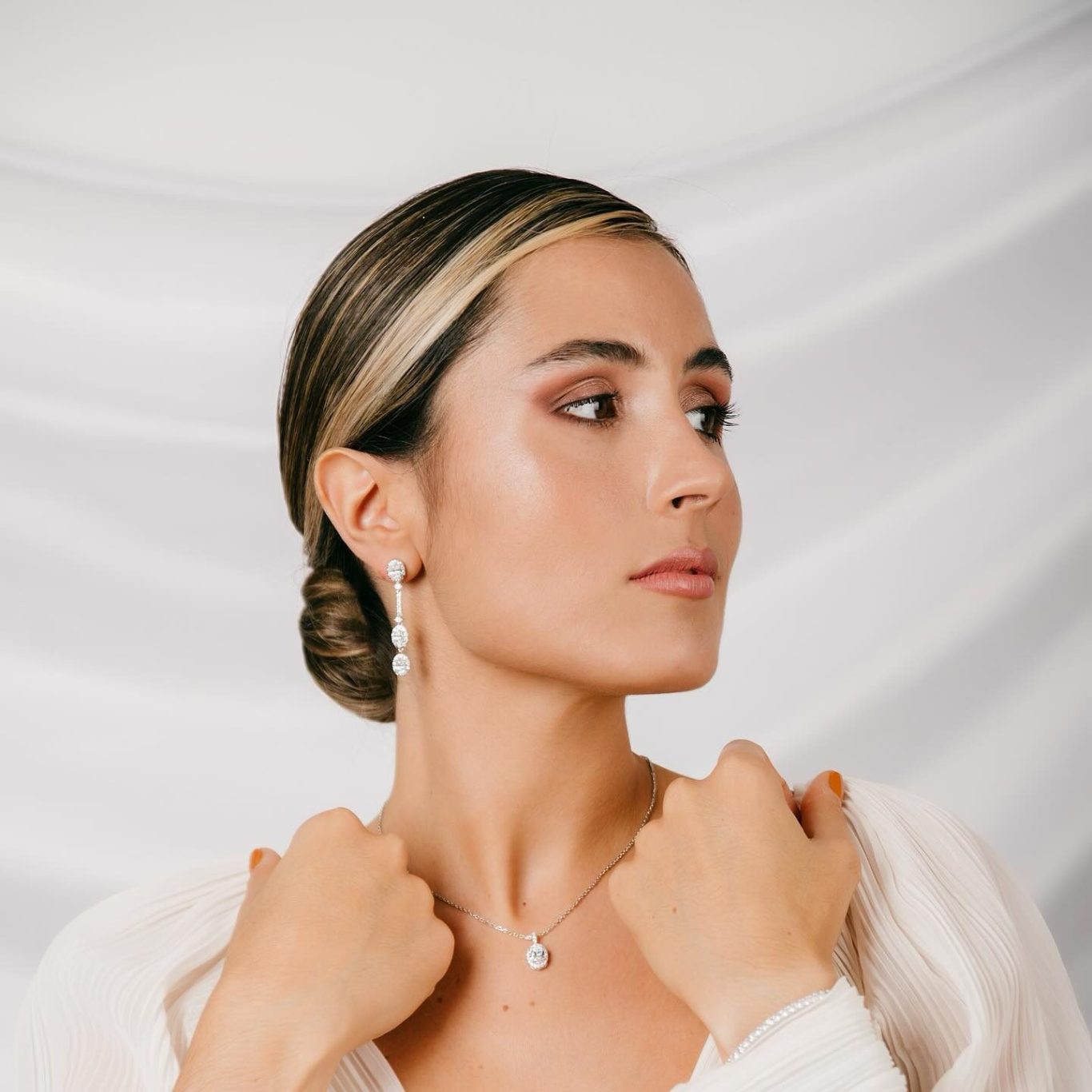 Mujer con peinado elegante, usando pendientes y collar, con fondo de tela clara.